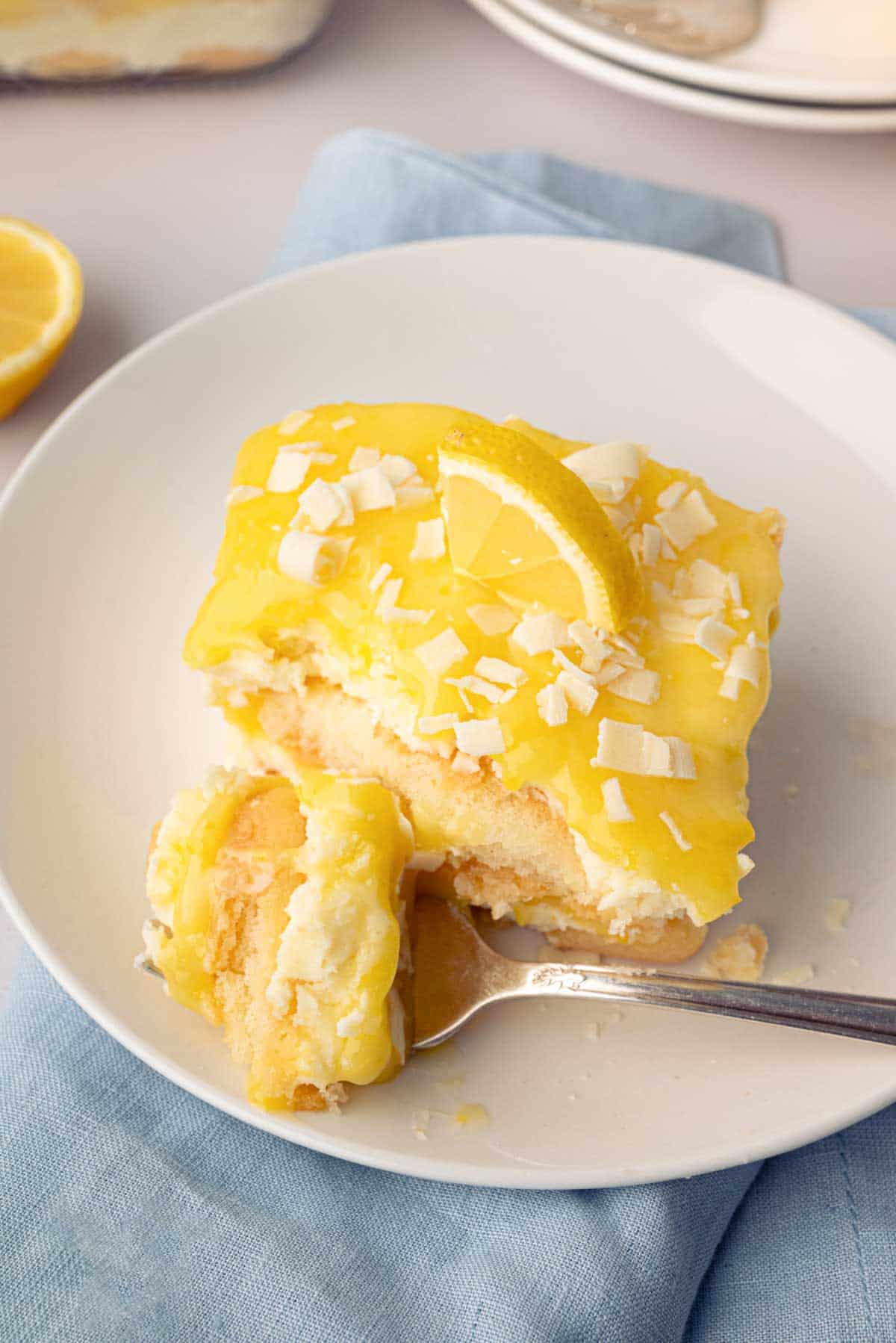 A slice of lemon tiramisu with a fork taking a bite out on a white plate with a blue napkin.