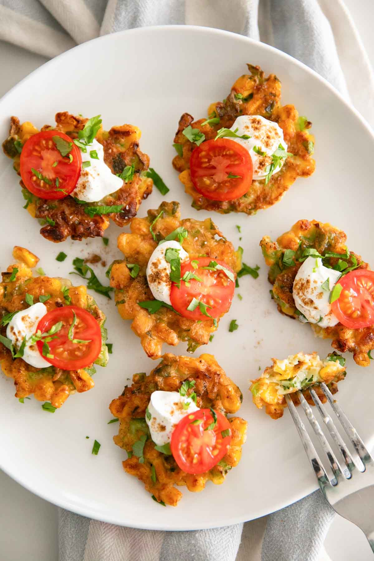 A white plate of corn fritters with cherry tomatoes and sour cream on top.