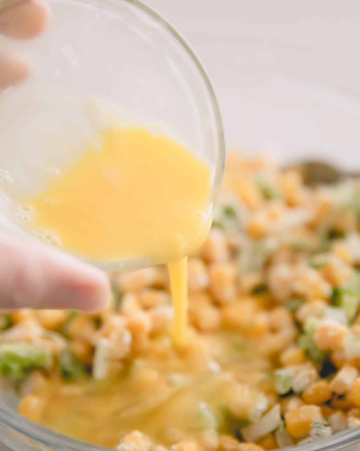 A hand pouring beaten egg from a glass bowl over corn fritter mixture.