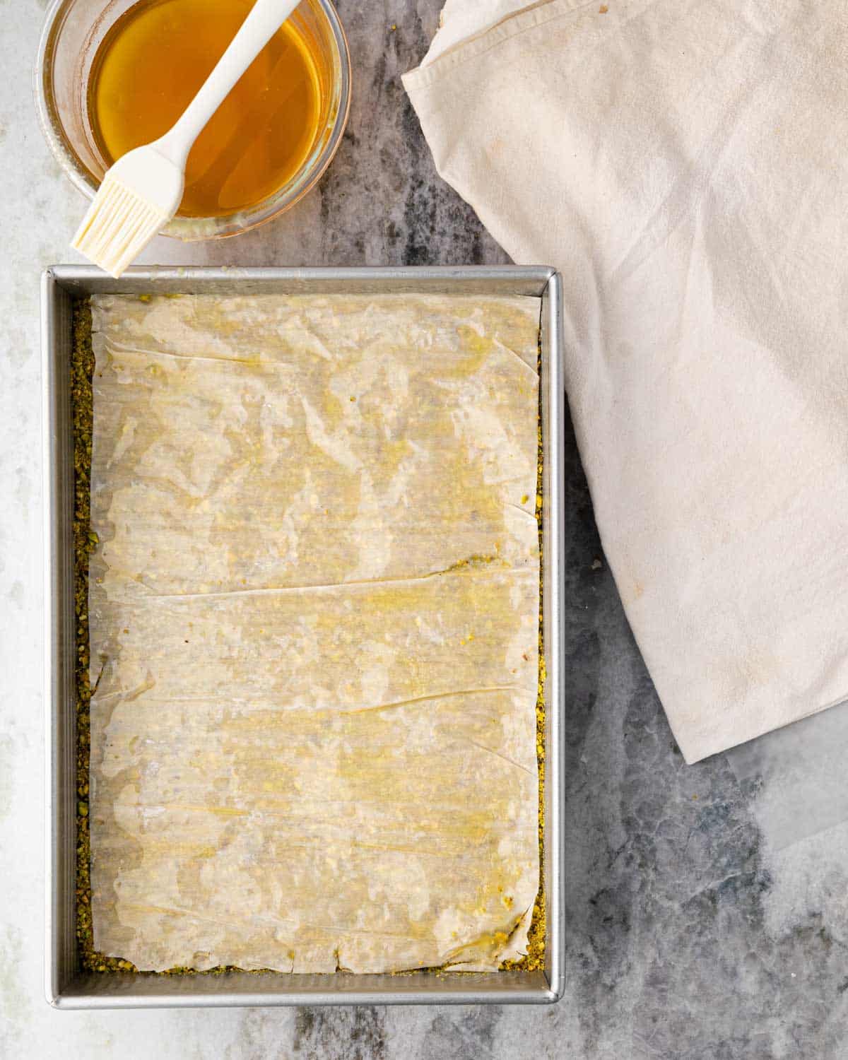 A baking dish with layers of pistachio, phyllo, and a bowl of simple syrup with a brush to the side.
