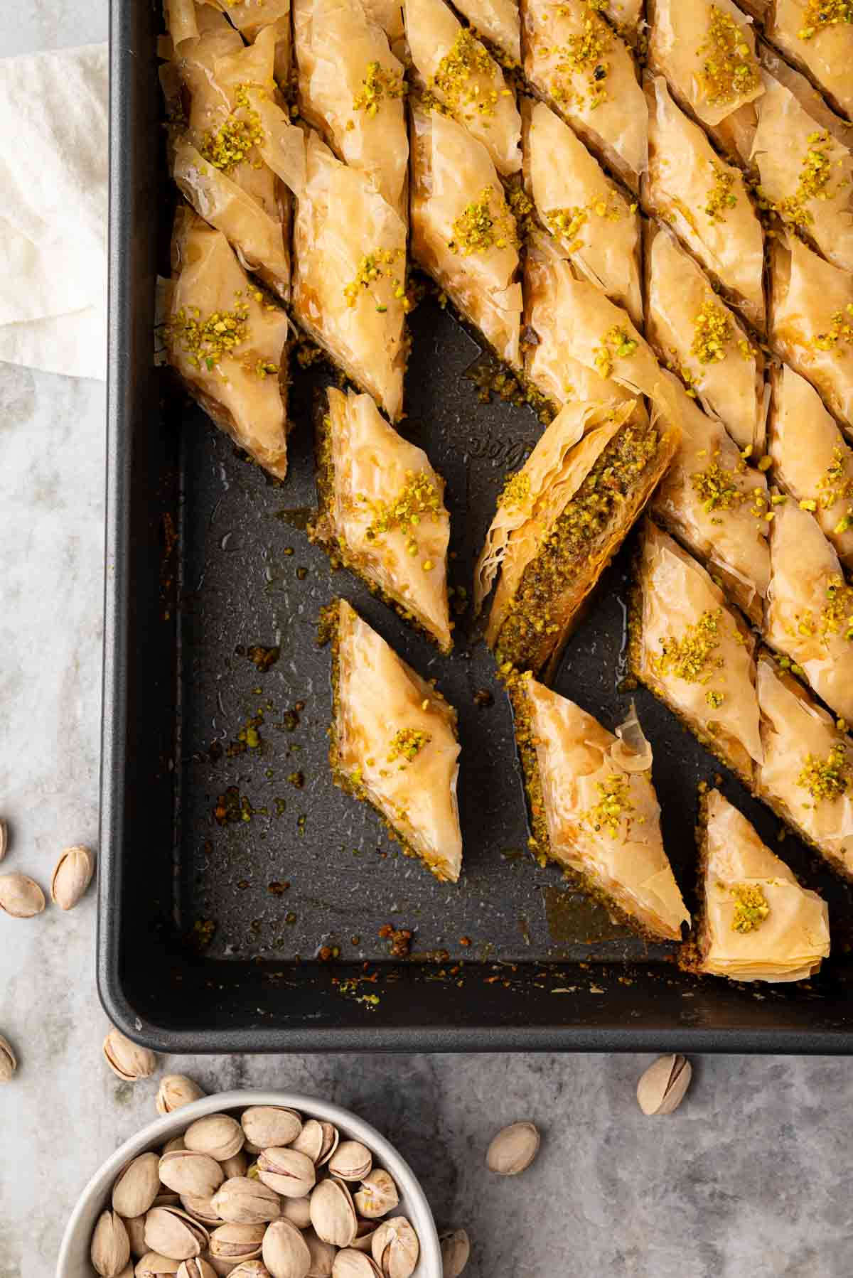 A baking dish with pistachio baklava with some pieces cut out and angled on their side. A bowl of pistachios is on the bottom.
