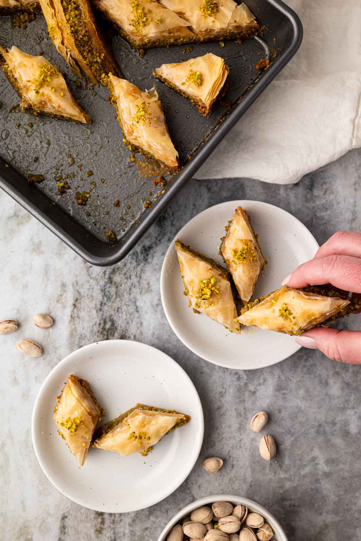 Two small white plates with pistachio baklava. A hand is grabbing a piece.