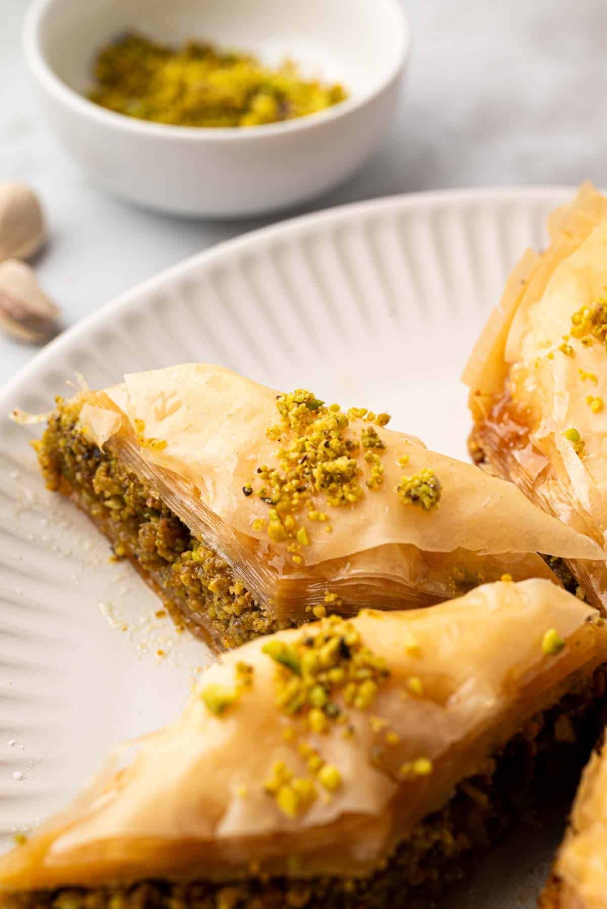 A piece of pistachio baklava on a plate with a bowl of chopped in the background.