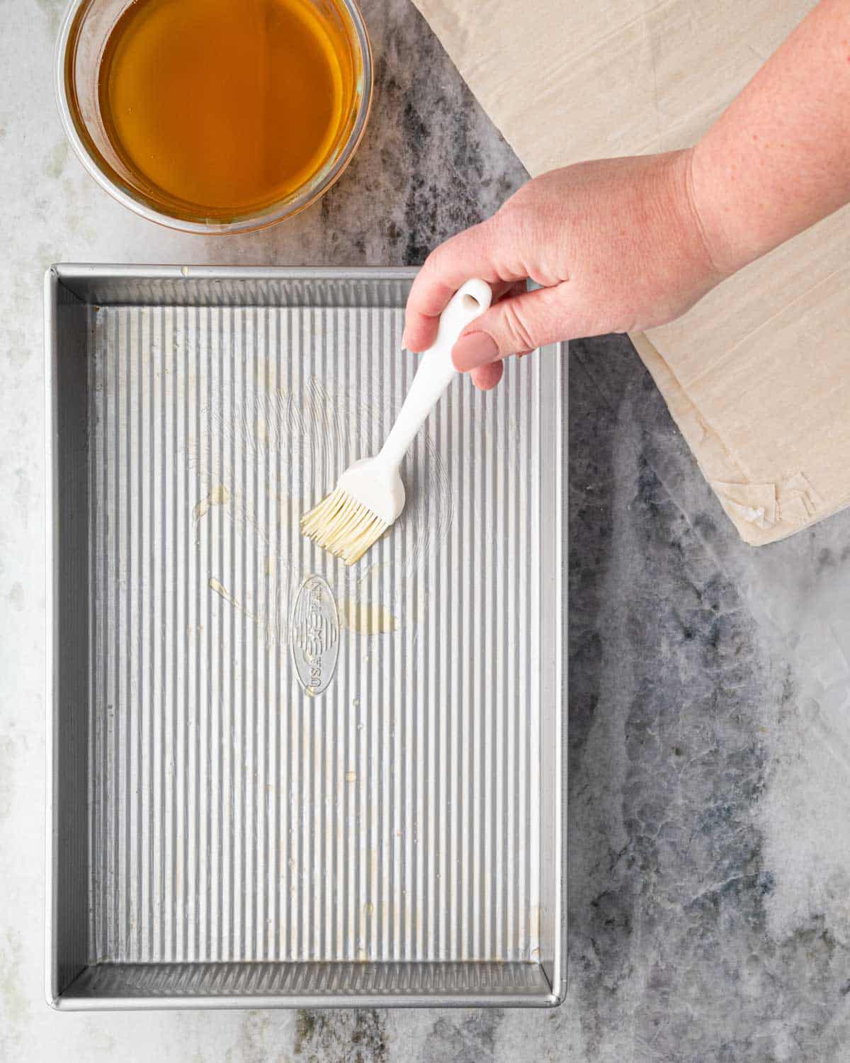 A hand brushing butter on the bottom of a baking dish. A bowl of simple syrup is to the side.