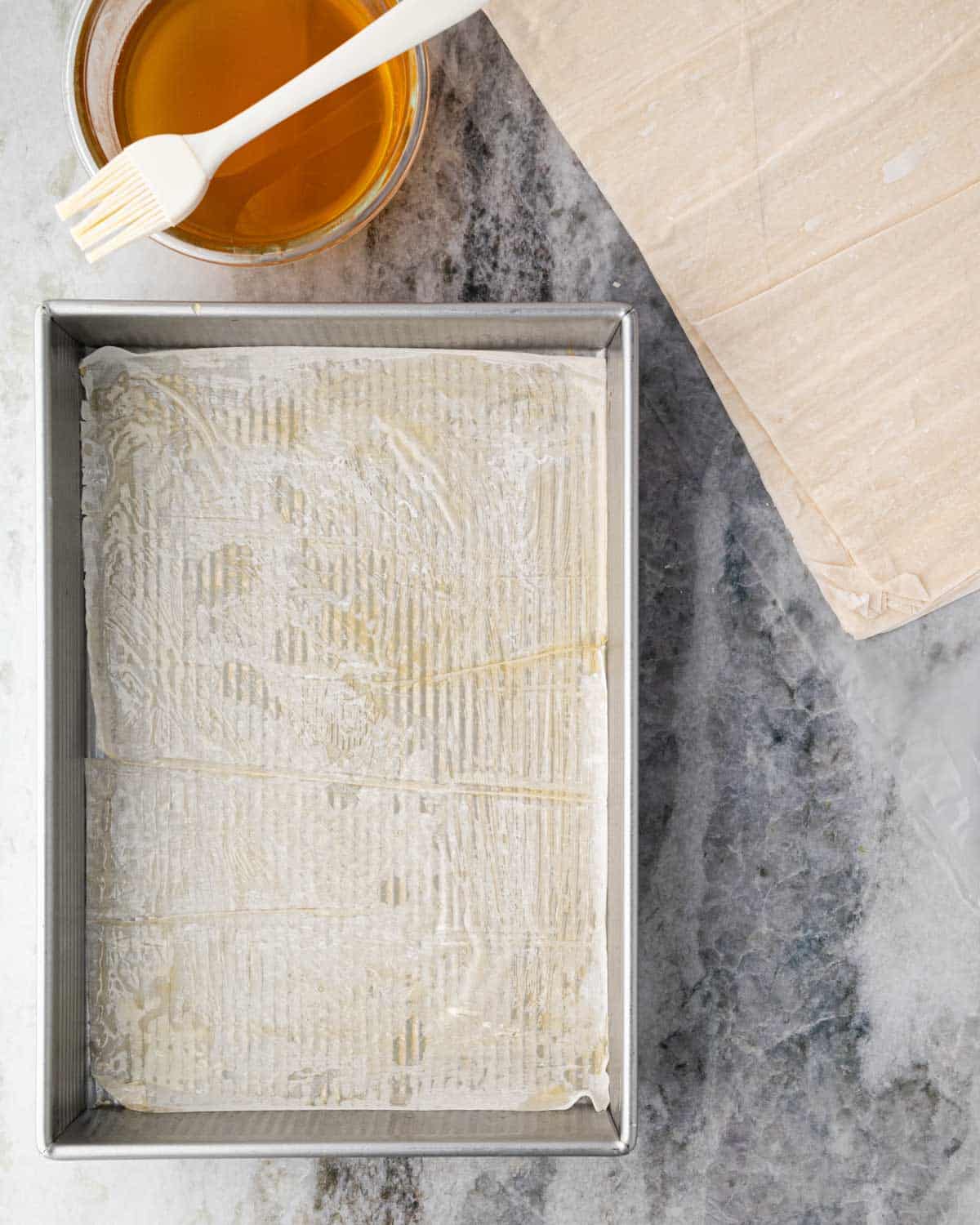 A metal baking dish with a sheet of phyllo on the bottom and clarified butter with a brush off to the side. Sheets of phyllo are next to the bowl of butter.