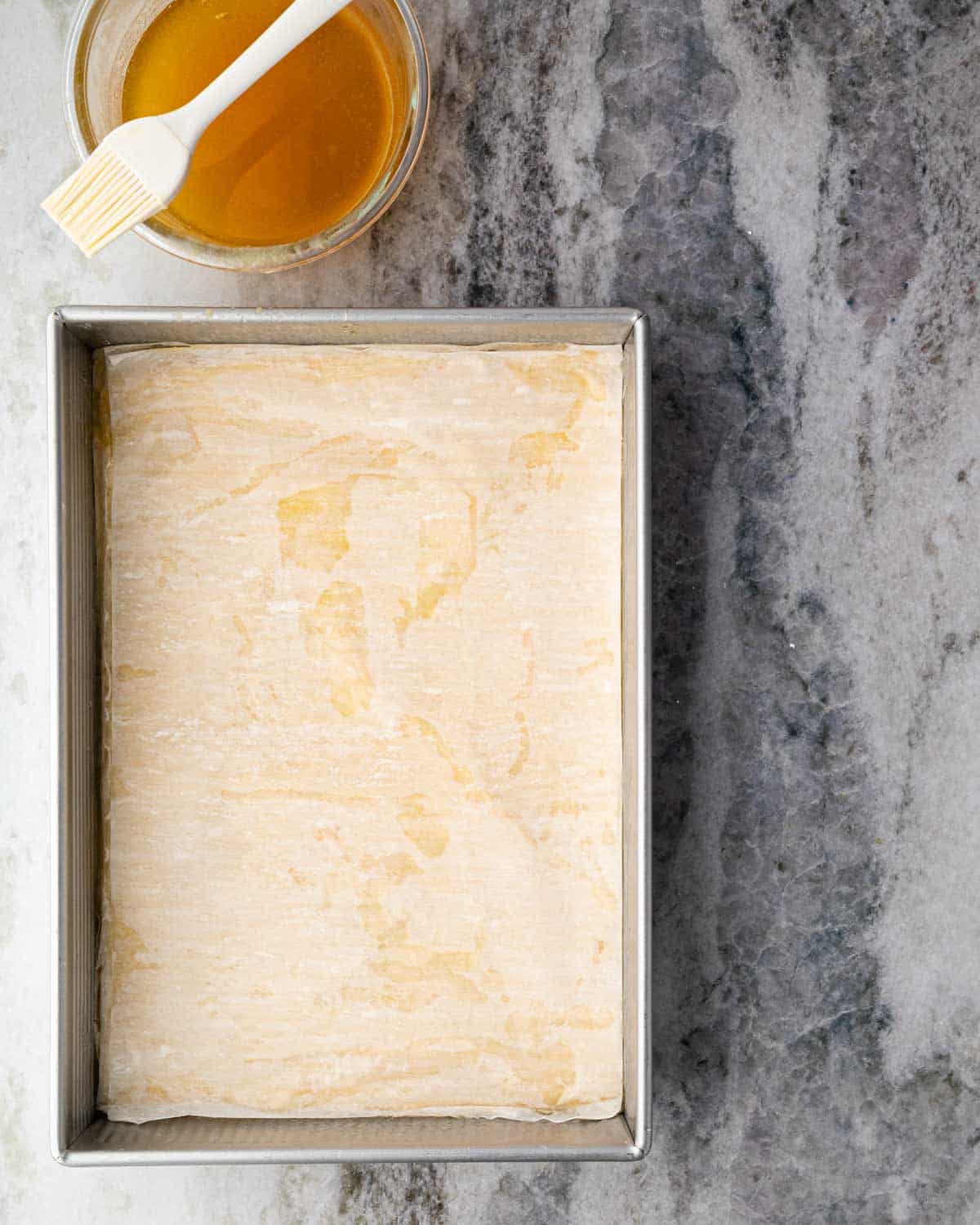 A baking dish with layers of phyllo brushed with butter and a nutty filling. A bowl of clarified butter with a brush is off to the side.