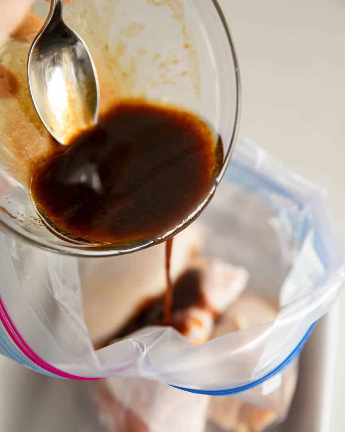 A glass bowl with a spoon pouring miso marinade into a ziptop bag with chicken thighs.
