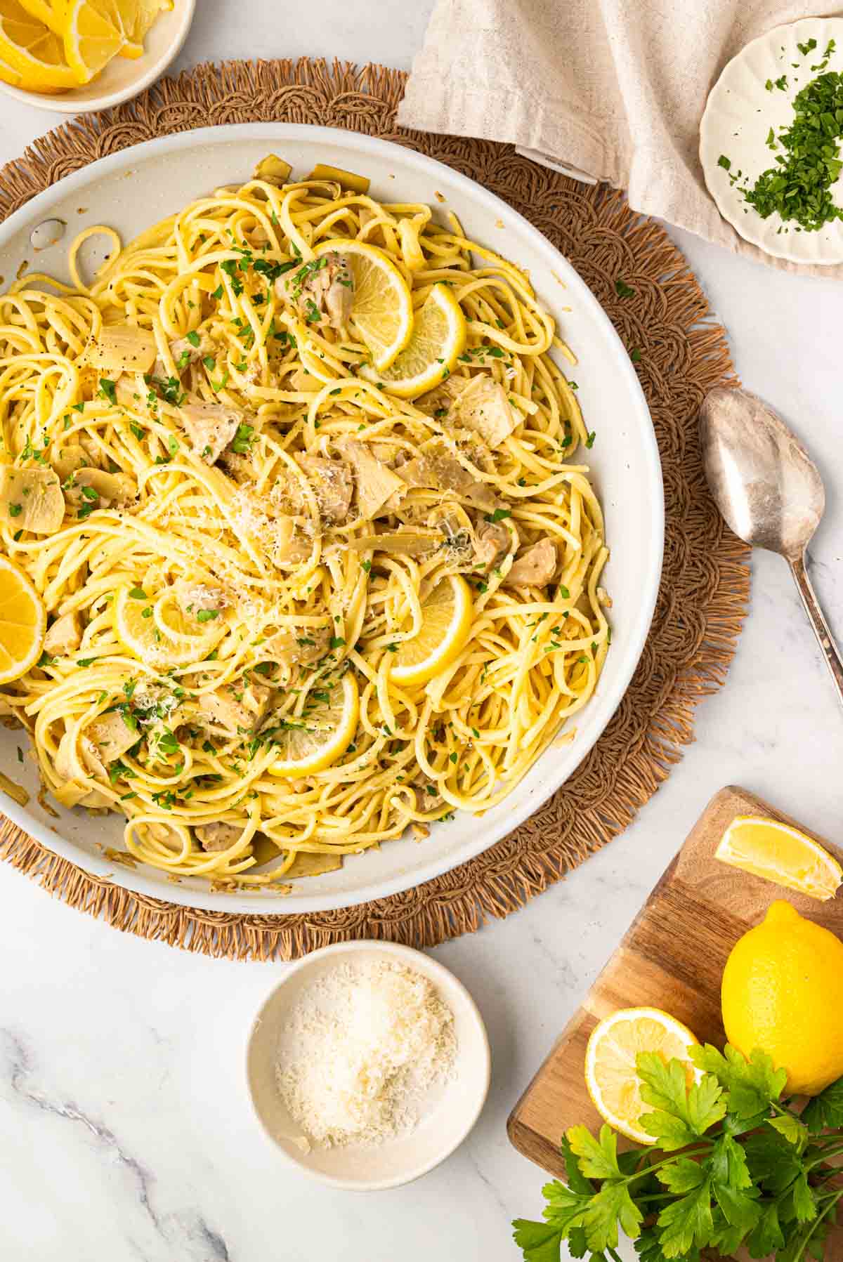 A white skillet with artichoke pasta on a wicker placemat with a spoon and grated parmesan cheese on the side.