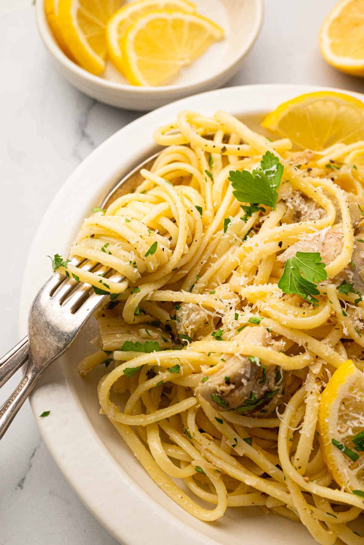 A close up image of lemon artichoke pasta with a fork and spoon in a white bowl.