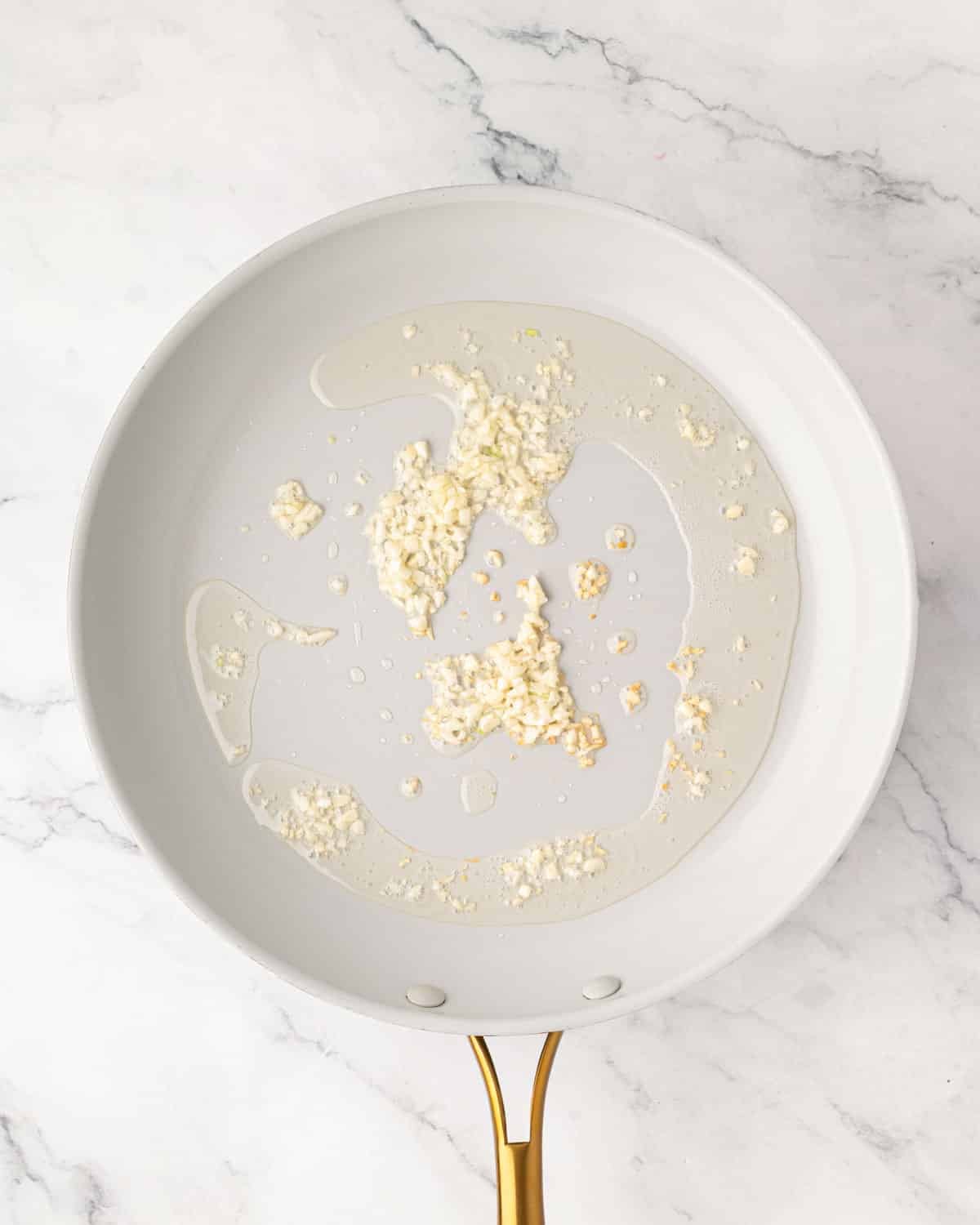 A white skillet with garlic cooking in oil.
