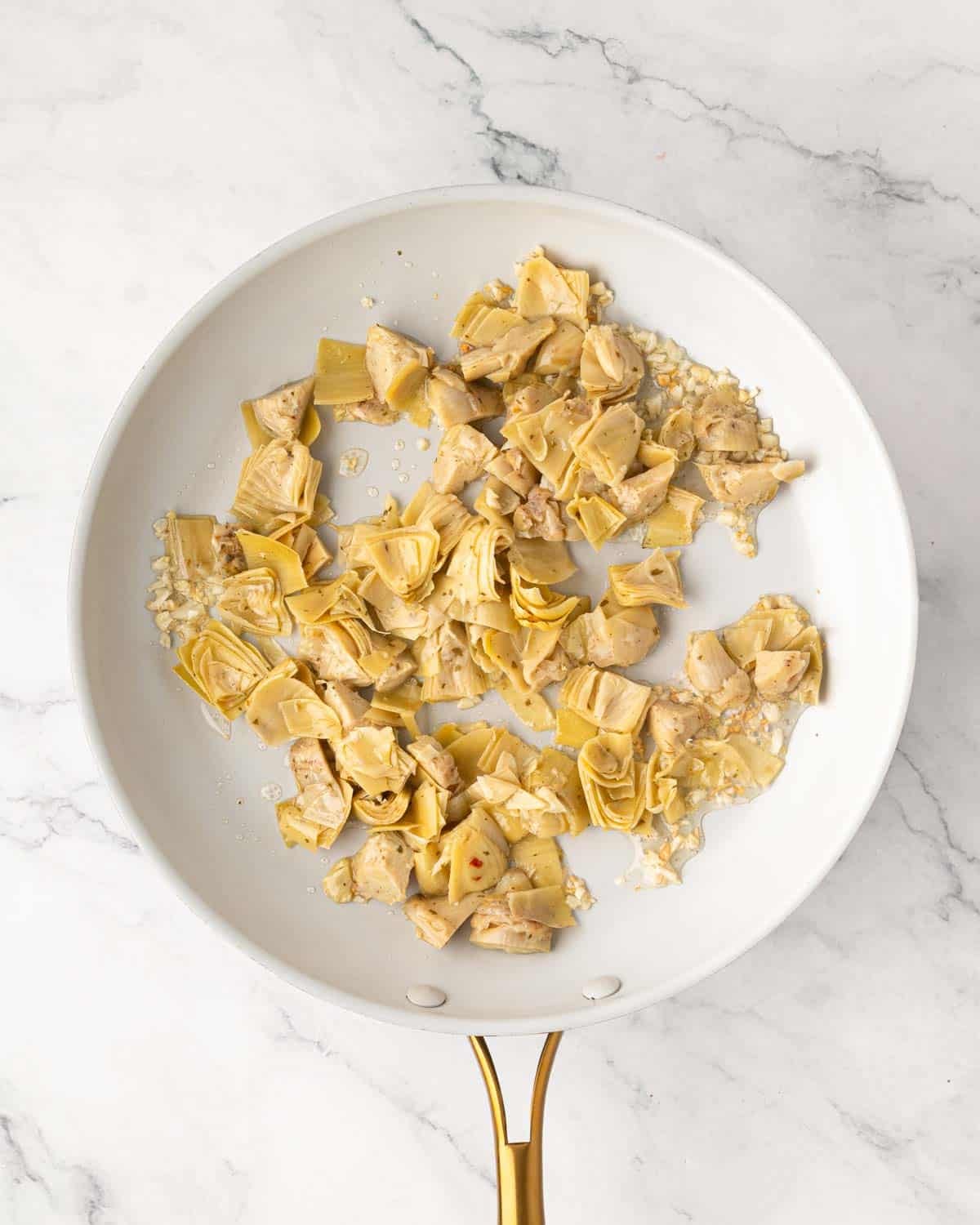 A white skillet with artichoke hearts nad garlic cooking.