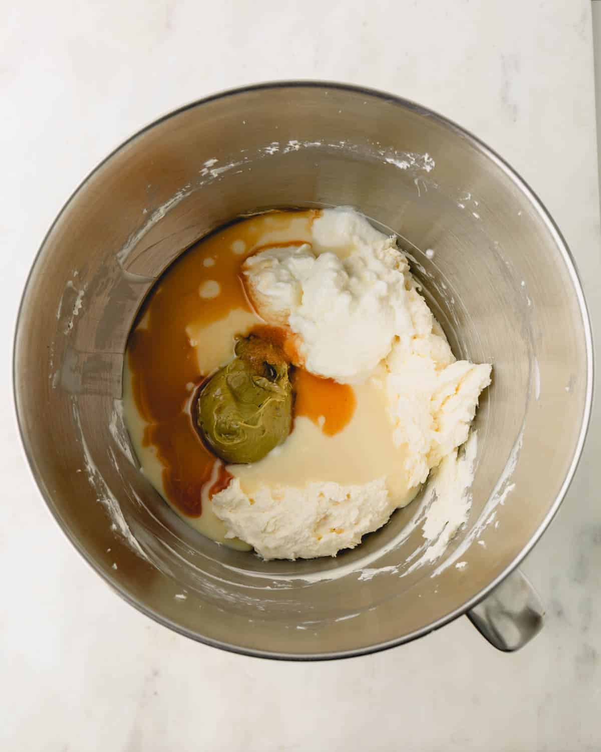 A stand mixer bowl with pistachio cheesecake filling ingredients inside. Pistachio butter and vanilla extract are on top.