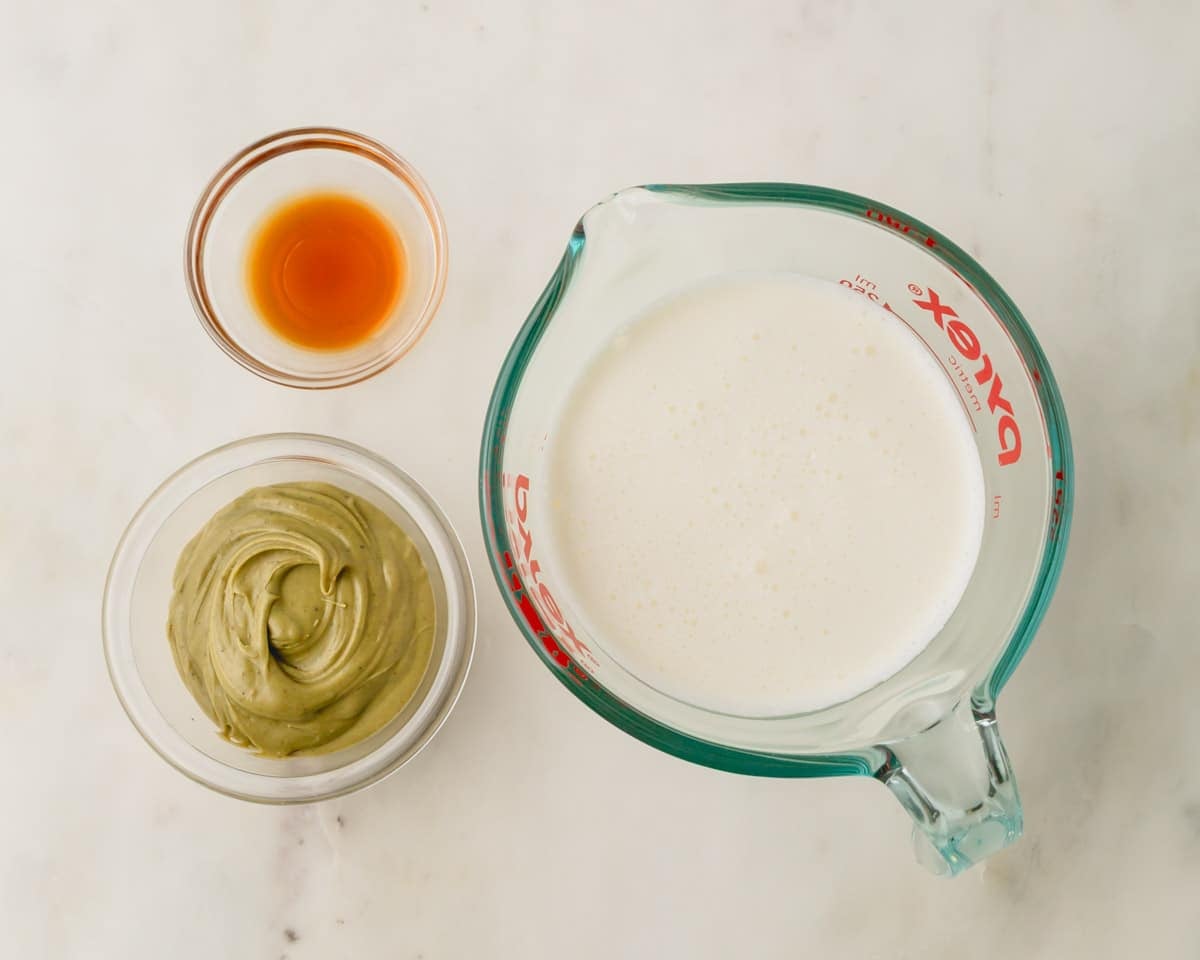 A bowl of pistachio butter, vanilla extract, and a measuring cup of heavy cream for pistachio whipped cream.