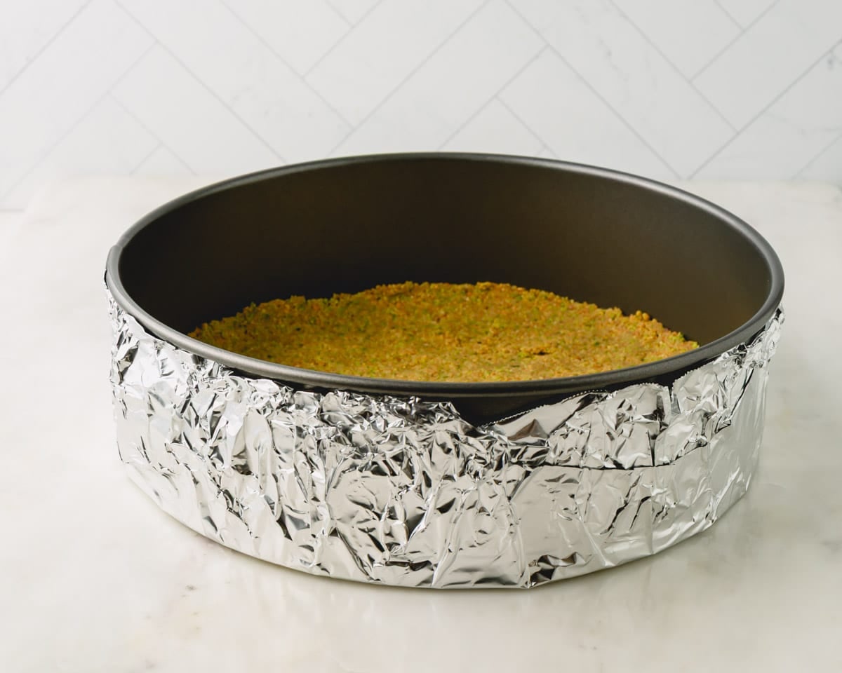 Pistachio graham cracker crust in a springform pan with foil wrapped around the bottom.