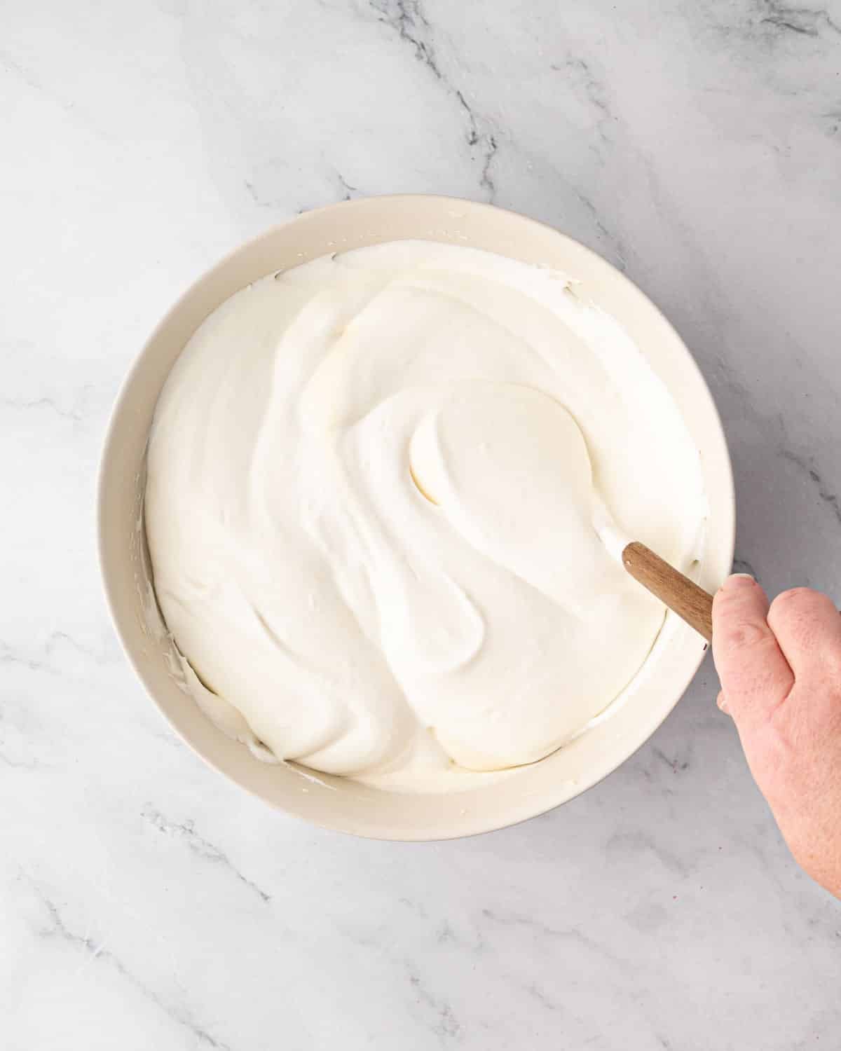 Whipped cream being folded into the whipped mascarpone with a spatula in a white bowl.
