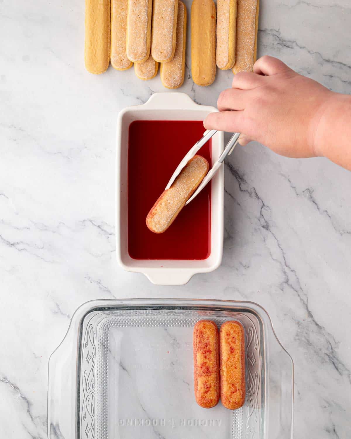 A hand dipping ladyfingers into raspberry simple syrup and placing them into the bottom of a glass baking dish.