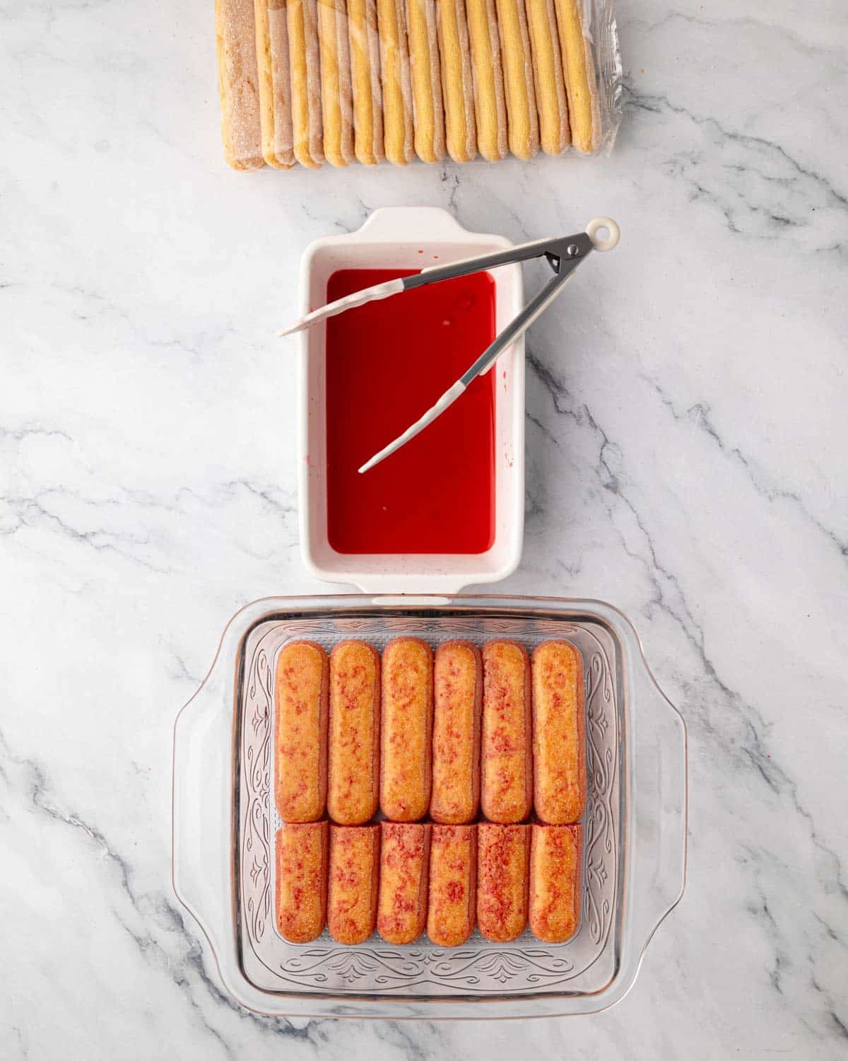 Ladyfingers are dipped in raspberry simple syrup in a small white dish and lined up in the bottom of a glass baking dish.