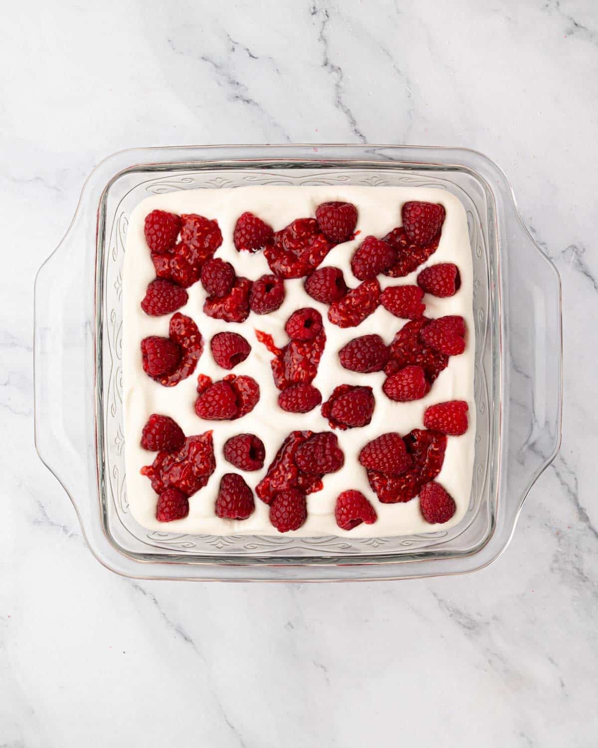 Raspberry compote and fresh raspberries are added on top of ladyfingers in a glass baking dish.