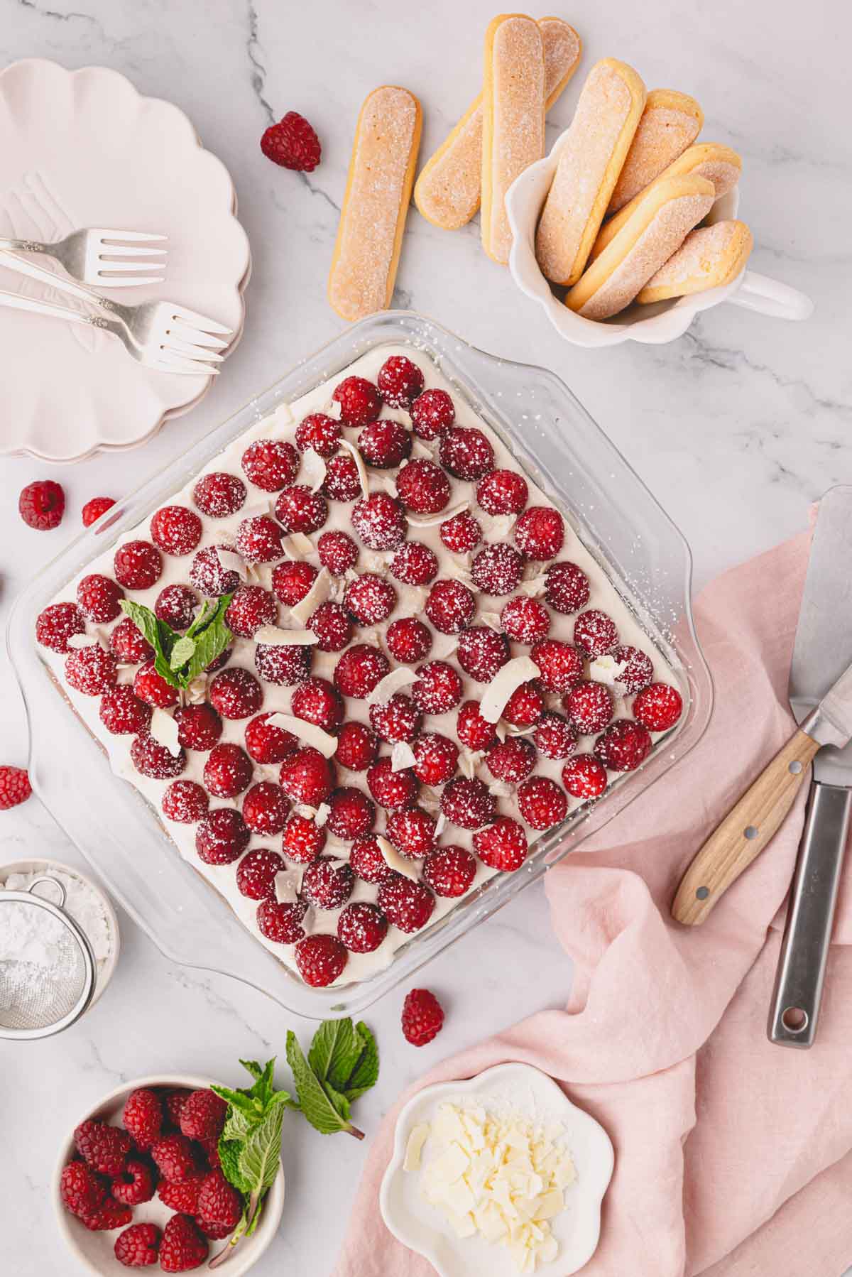 Finished raspberry tiramisu with fresh raspberries on top and plates and fresh raspberries to the side.