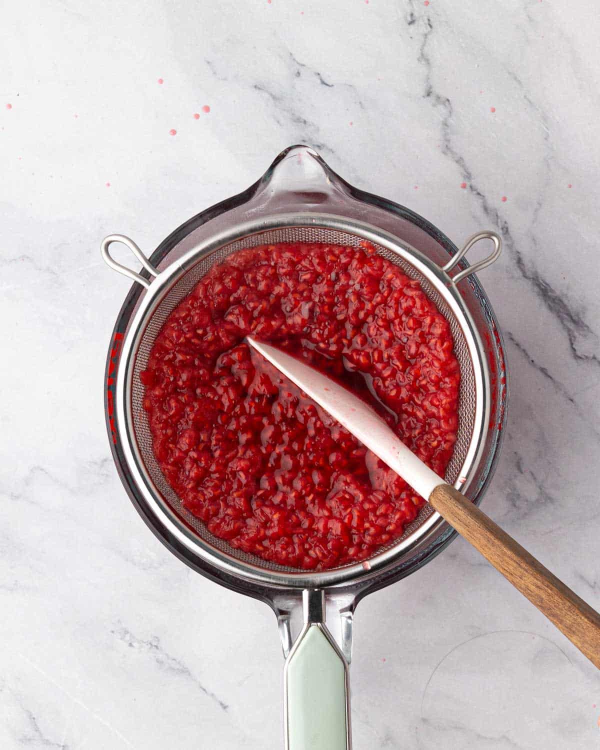Straining raspberry simple syrup in a mesh sieve over a glass measuring up. A white spatula is resting on top.
