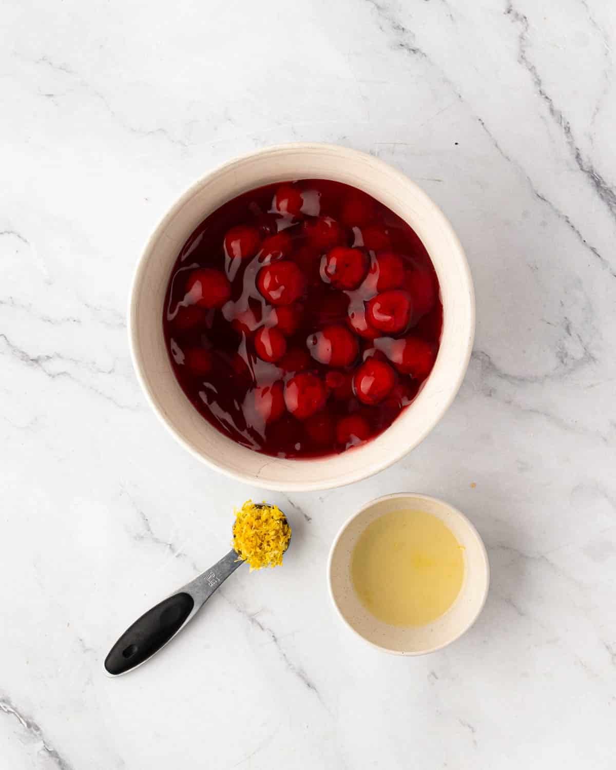 A white bowl with cherry pie filling, a measuring spoon with lemon zest, and lemon juice in a small white bowl.