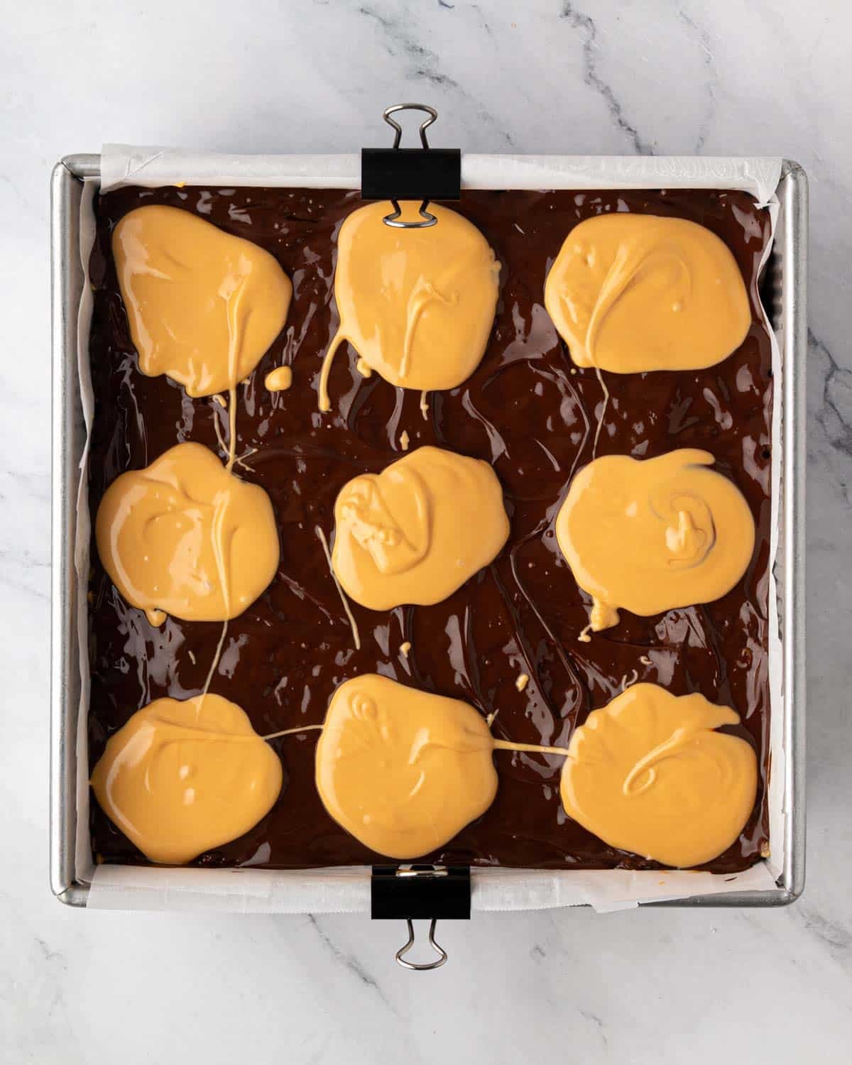 A baking pan with scotcharoos, melted chocolate is dolloped with melted butterscotch for topping.