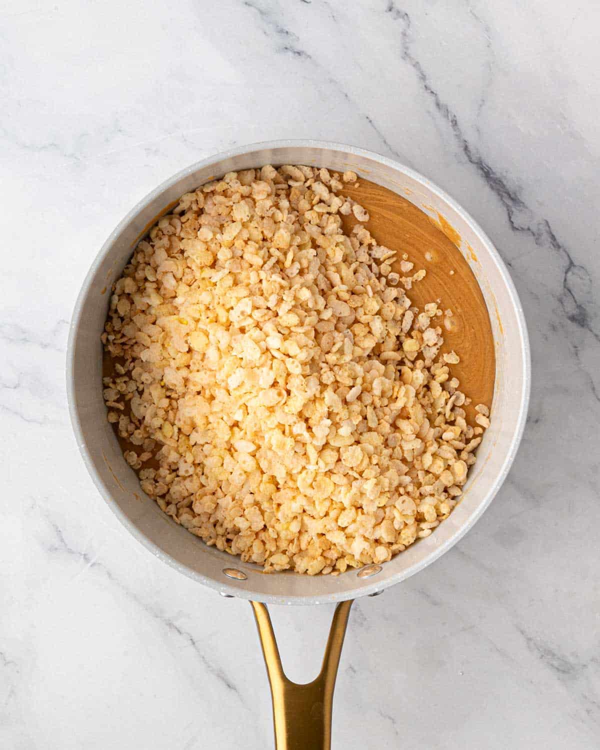 A white saucepan of melted peanut butter mixture with rice crispy cereal for scotcharoo base.