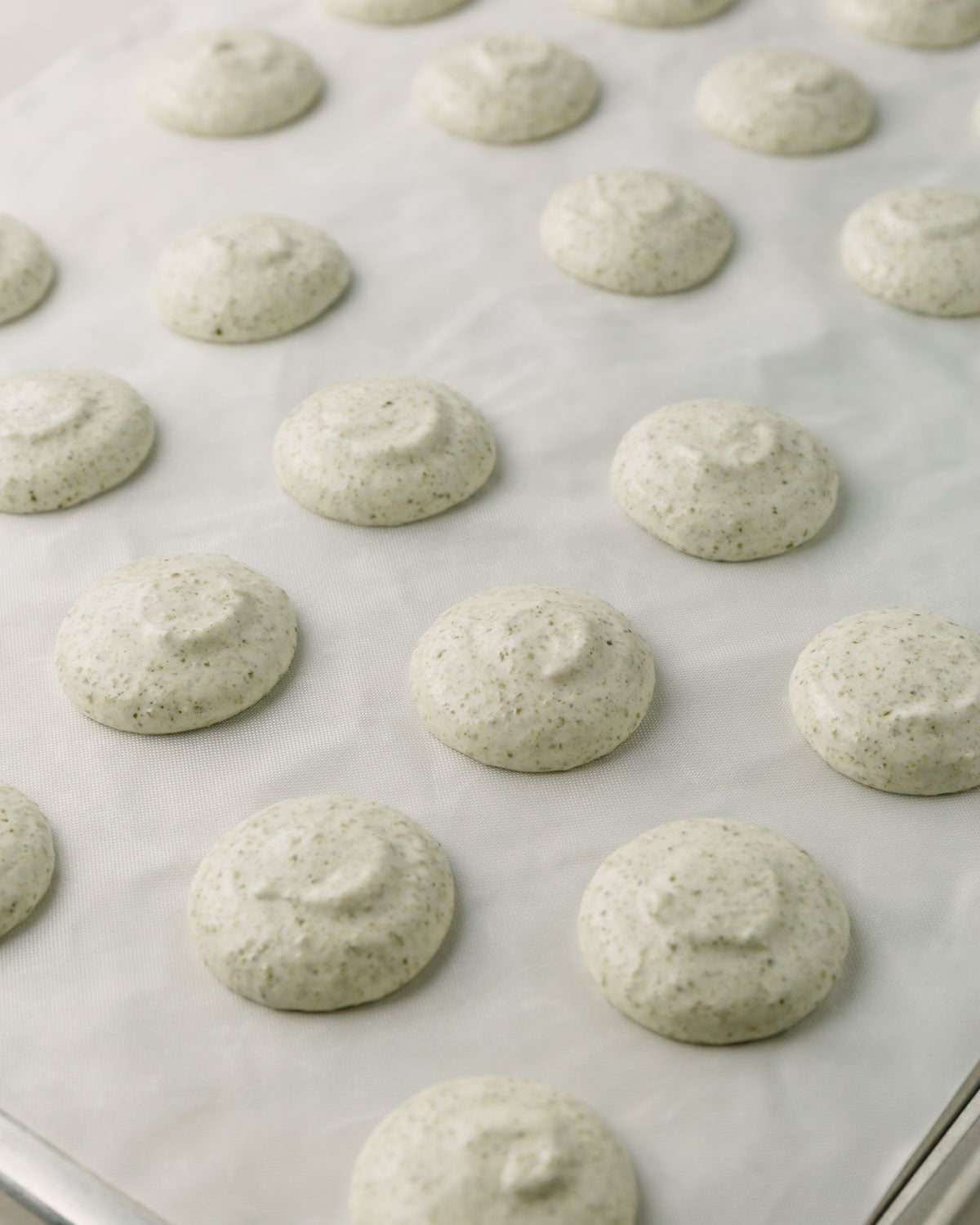 Piped macaron shells on baking sheet lined with white teflon sheet.